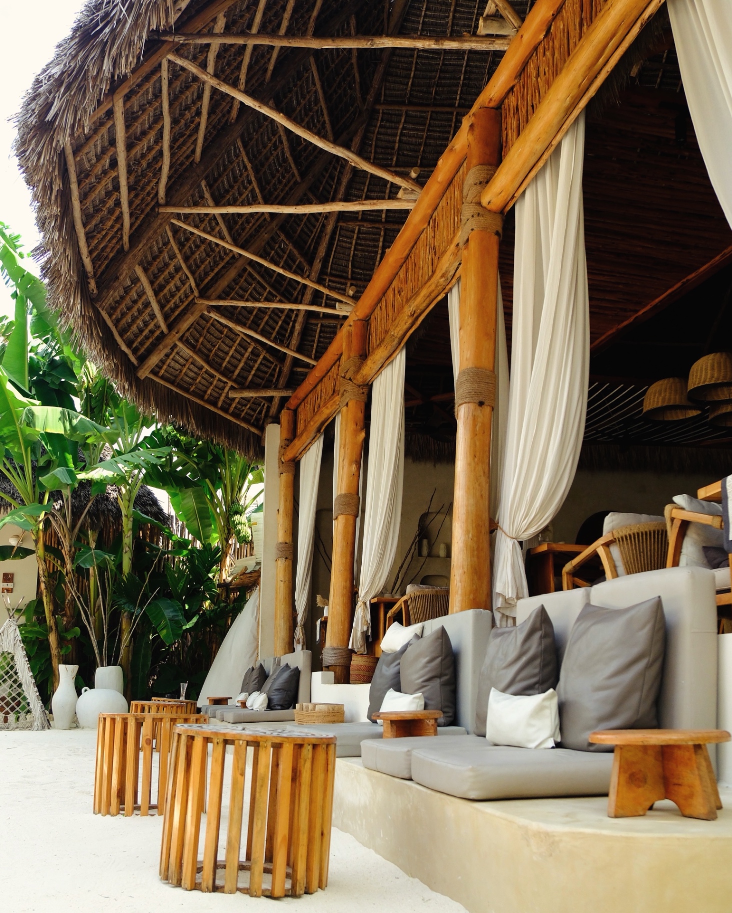 Open-air makuti-roofed lounge at Kai Zanzibar with timber columns, linen curtains and sand floor