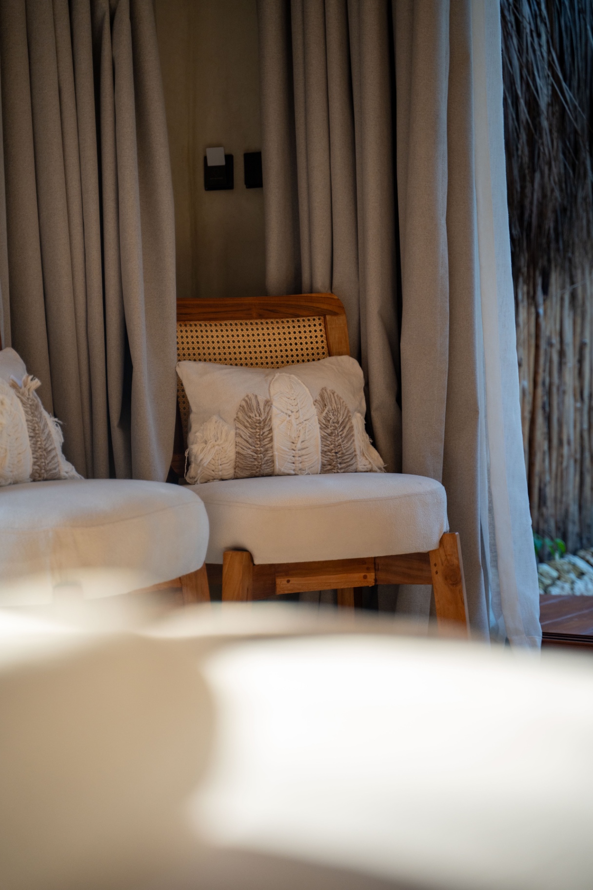 Cane and teak armchair with a tufted cushion behind linen curtains